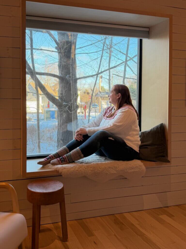 Woman sitting on the window of a cabin looking out at a snowy scene.