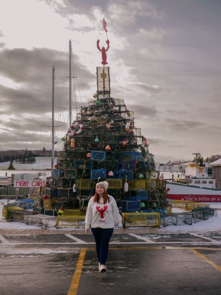 Woman wearing a lobster Christmas jumper stands in front of a lobster trap Christmas tree in Lunenburg, Nova Scotia.