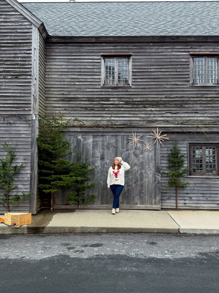 Woman wearing a lobster Christmas jumper that says Santa Claws stands in front of a Christmas tree at a rustic barrel factory in Shelburne, Nova Scotia