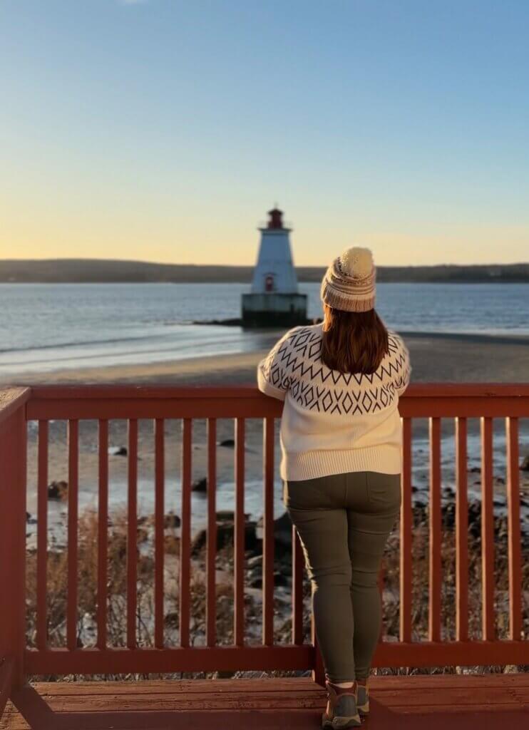 Woman wearing a cream knitted jumper and hat looks out at Sandy Point lighthouse in Nova Scotia.