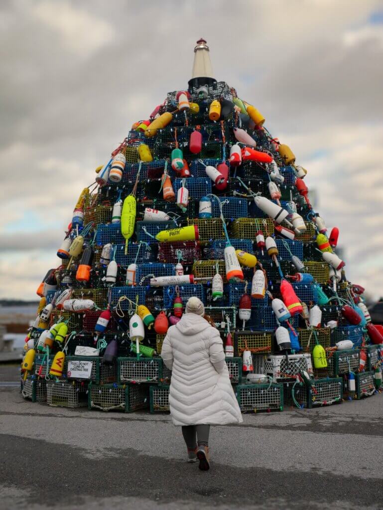 Woman wearing a padded white coat stands in front of the lobster trap Christmas tree at Cape Sable Island near Barrington, Nova Scotia.