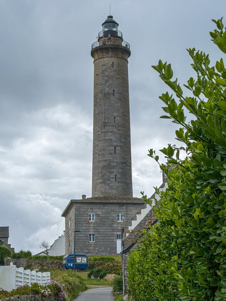 A low-angle view of the tall stone Phare Île de Batz lighthouse rising from a square base under an overcast, cloudy sky, with a narrow paved pathway leading towards it, bordered by lush green bushes and a white fence.