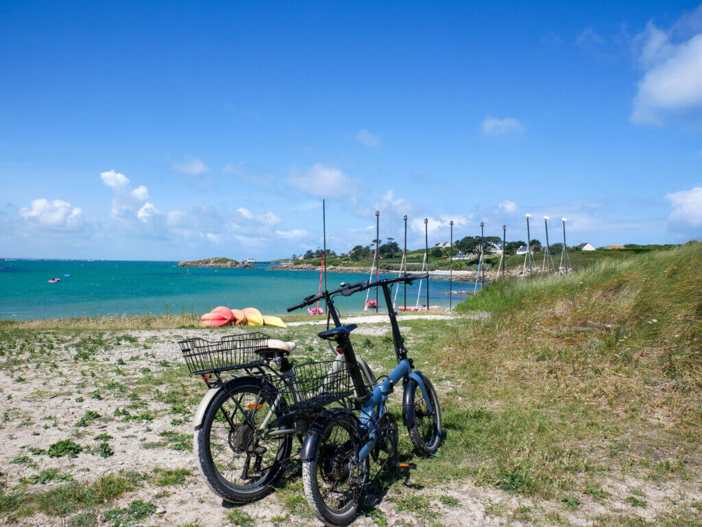 Two rental e-bikes with black luggage baskets parked on a grassy, sandy coastal path on Île de Batz, overlooking a vibrant turquoise sea, a distant sandy cove, and a bright blue sky with scattered white clouds.