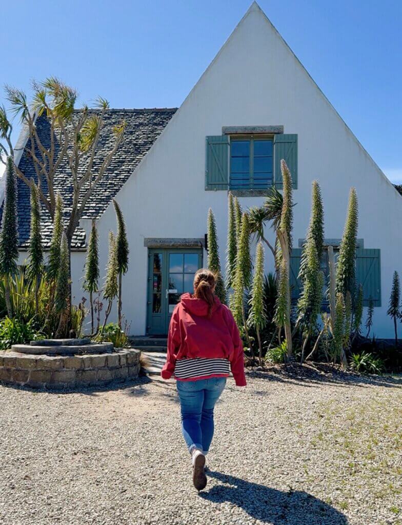 Rear view of a woman in a red jacket walking along a gravel path towrds the entrance to the Jardins Georges Delaselle on Île de Batz, framed by tall spiky Echium plants, palm trees, and sage-green window shutters under a clear blue sky.