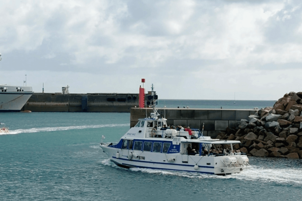 A blue and white passenger ferry boat carrying travellers through turquoise waters, navigating out of a harbour with a stone breakwater under a cloudy sky on the way to Île de Batz.