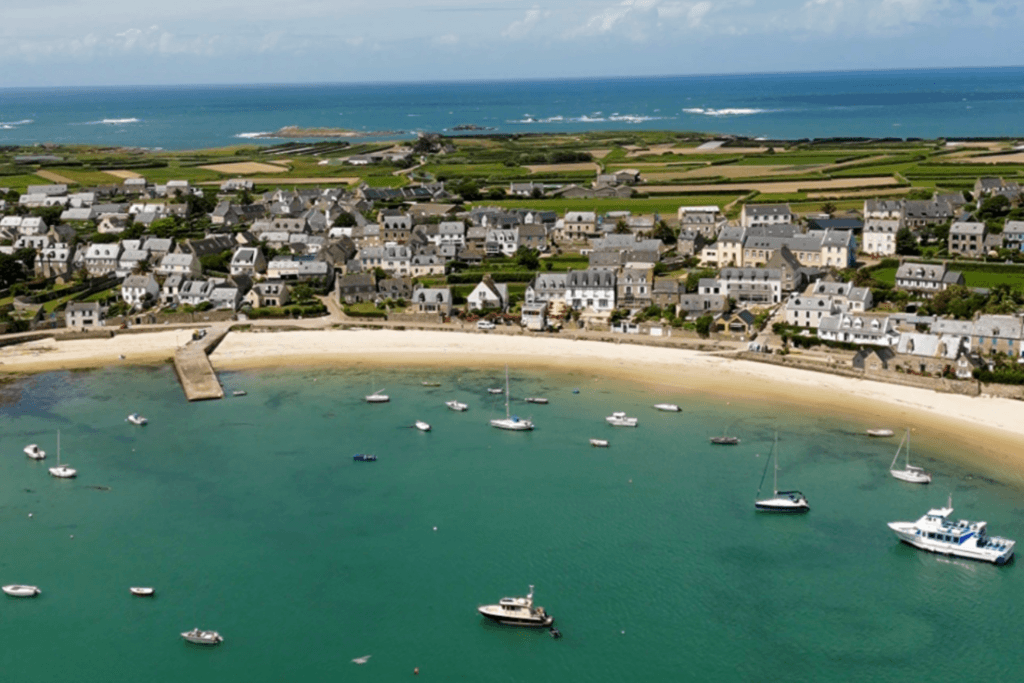 Aerial view of the harbour town on Île de Batz, showing a cresent-shaped white sand baech meeting clear turquoise water filled with scattered boats, lined with traditional stone houses and a patchwork of green argicultural fields extending to the Atlantic ocean.