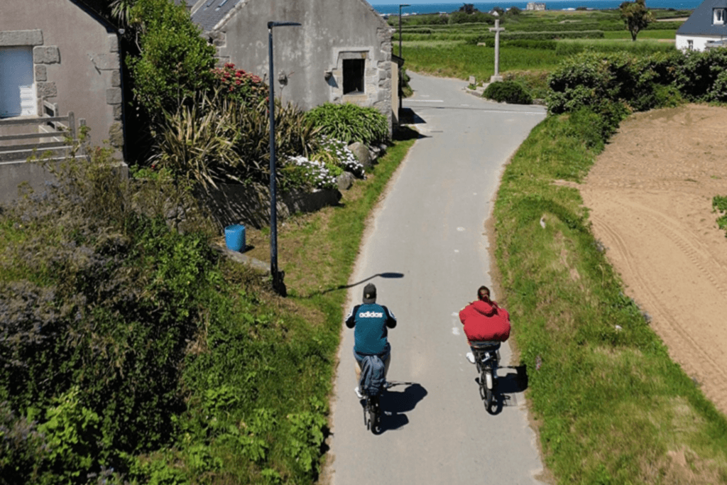 An elevated, rear view of two people cycling down a quiet asphalt road on Île de Batz, surrounded by green grassy banks, traditional stone houses with gardens, and rural fields stretching out towards the sea in the distance.