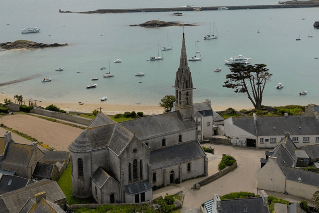 A high-angle view of a traditional stone church with a tall, pointed spire on Île de Batz, overlooking a sandy beach and a calm bay dotted with numerous small boats and sailing yachts under an overcast sky.