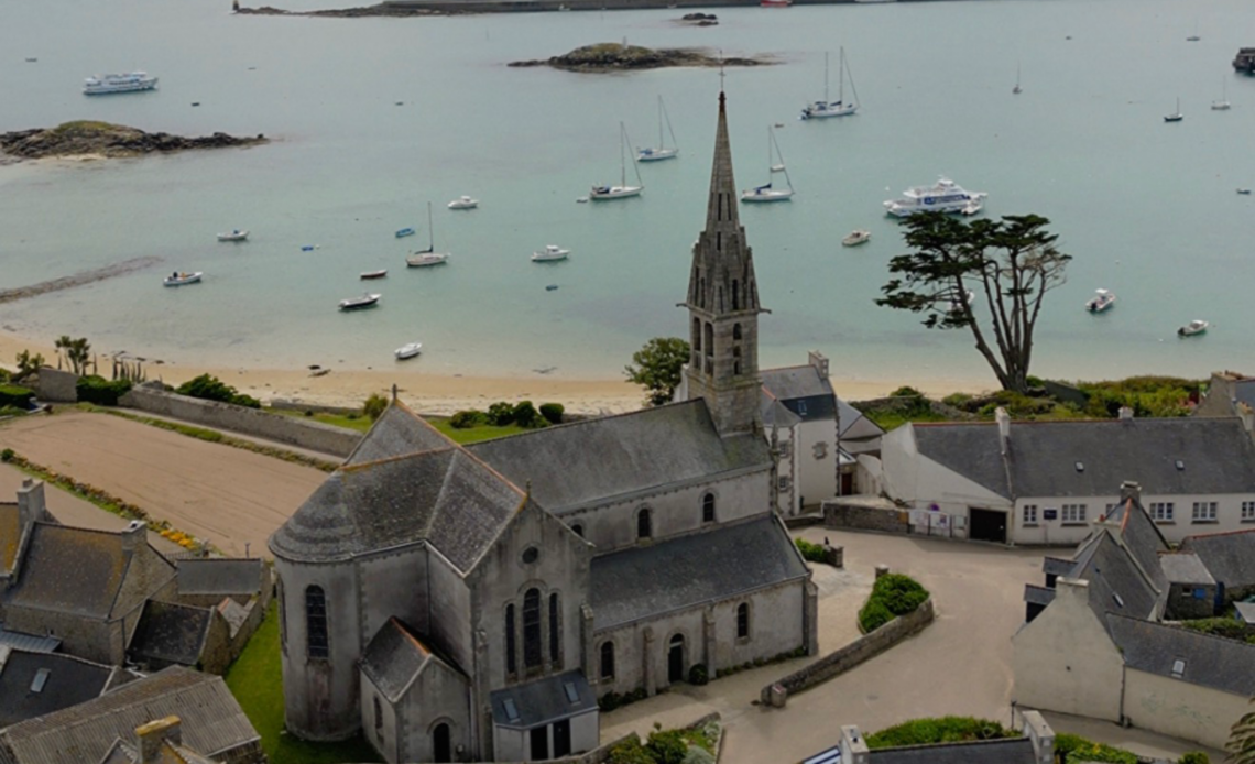 A high-angle view of a traditional stone church with a tall, pointed spire on Île de Batz, overlooking a sandy beach and a calm bay dotted with numerous small boats and sailing yachts under an overcast sky.
