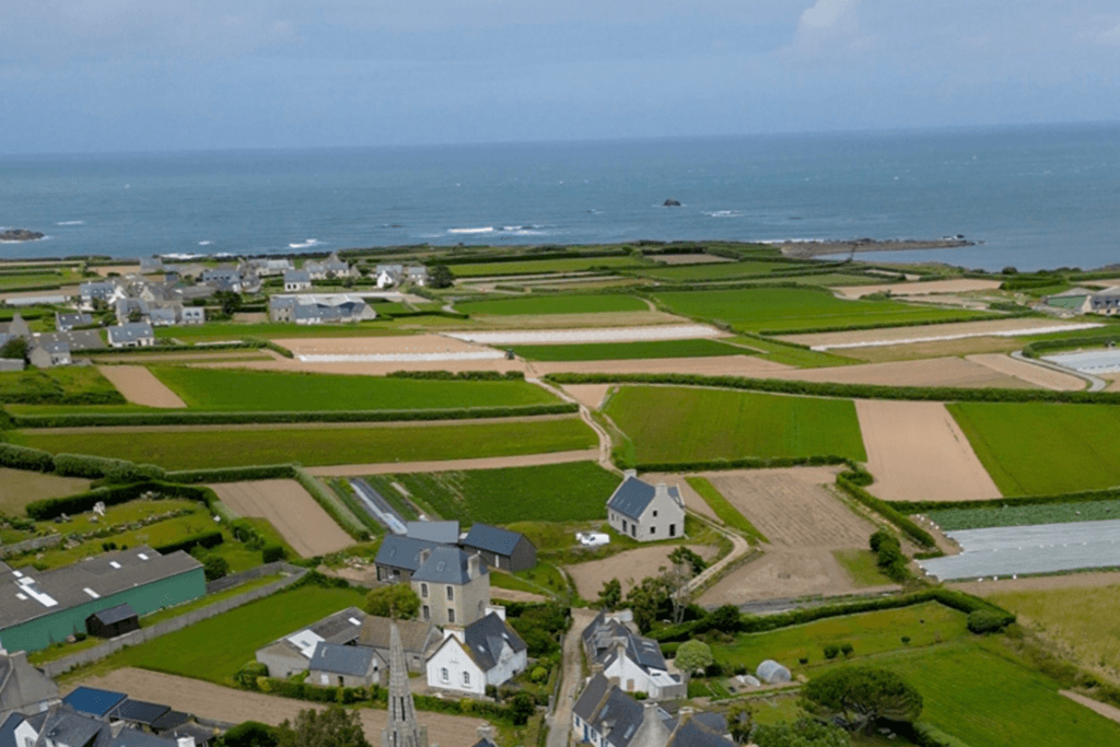 A high-angle, panoramic view looking out over the patchwork landscape of Île de Batz, featuring geometric green and brown agricultural fields, scattered coastal stone houses, a church spire in the lower foreground, and the deep blue sea stretching to the horizon.