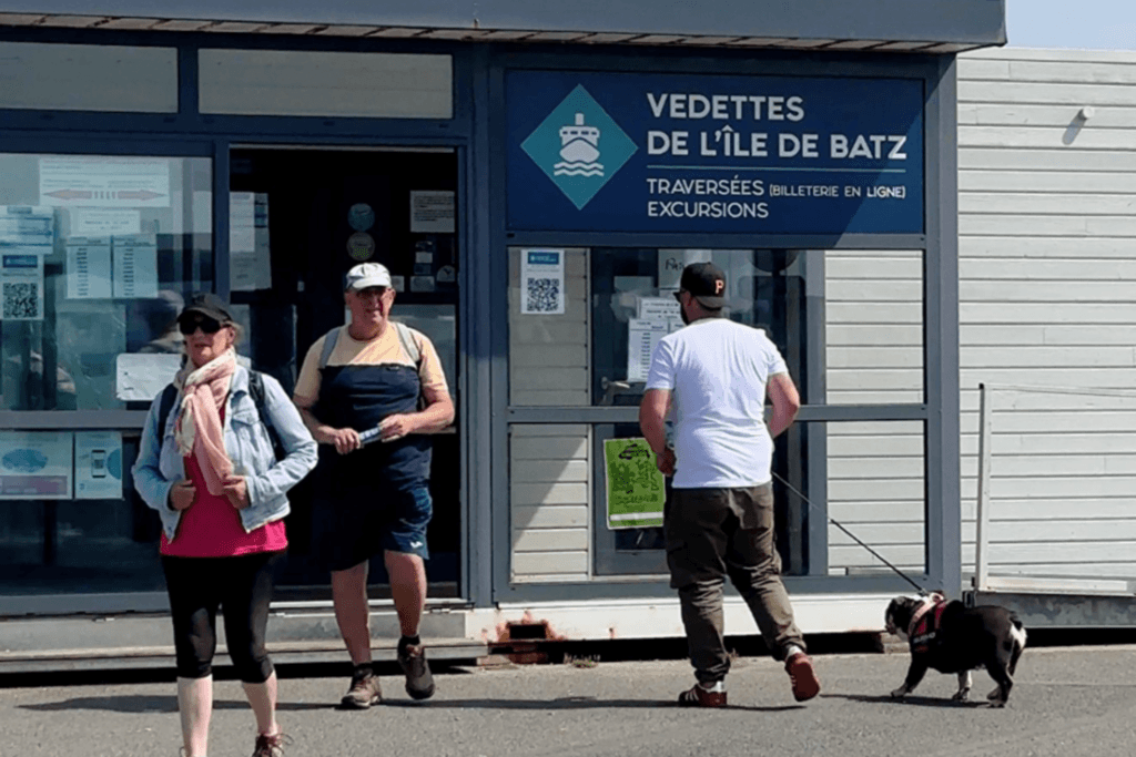 A couple, walking past the "Vendettes de l'Île de Batz" ticket office, and a man walking his black dog on a leash in front of them, in front of the ticket office building. This photograph looks straight at the building in Roscoff, which has white wooden paneling and a dark-framed glass front, with "VEDETTES DE L'ÎLE DE BATZ" and "TRAVERSÉES (BILLETTERIE EN LIGNE) EXCURSIONS" clearly displayed on a blue sign. A man in a white t-shirt and khaki pants stands in front of the office, his black english bulldog on a pink leash. The building sits on a sun-drenched gravel and asphalt lot near the harbour. The scence is bright with strong daylight and well-defined shadows, capturing the textures of the old buildings and the activity of the ferry passengers.
