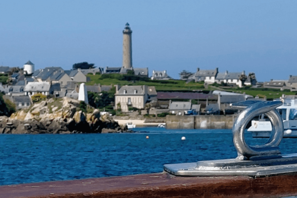 A view from a boat looking across the blue sea toward the village of Île de Batz, featuring a silver metal mooring cleat on the wooden deck in the foreground, with coastal stone houses, a small harbour, and the iconic tall stone lighthouse rising under a clear blue sky.