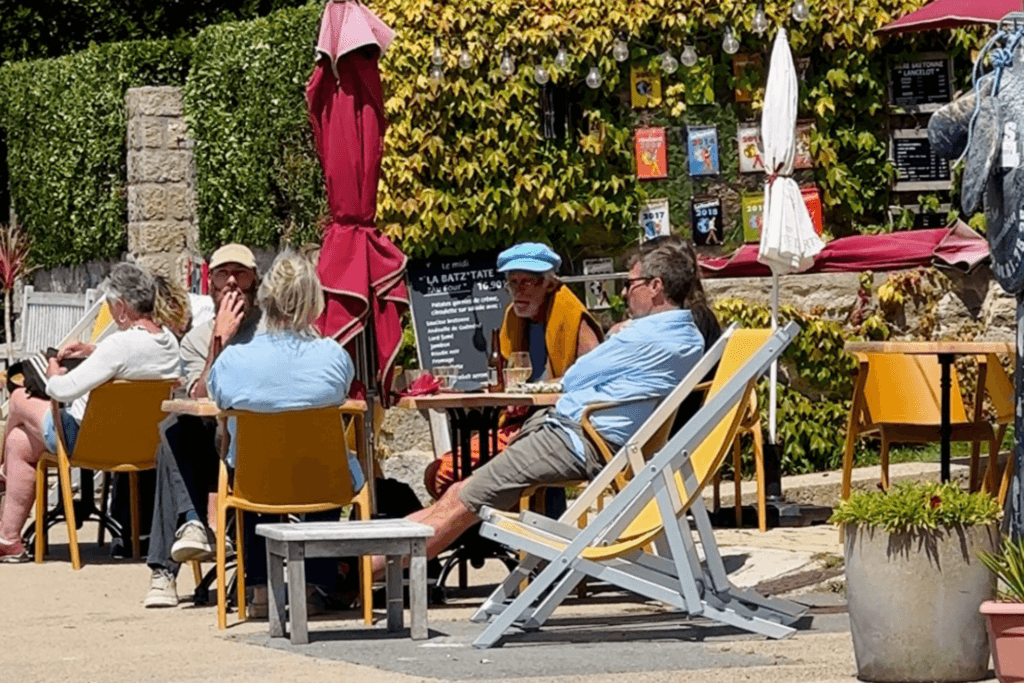Patrons relaxing at outdoor tables on a sunny terrace at a  café on Île de Batz, featuring people chatting in deckchairs and yellow seats surrounded by lush green foliage, string lights, and a chalkboard menu.      