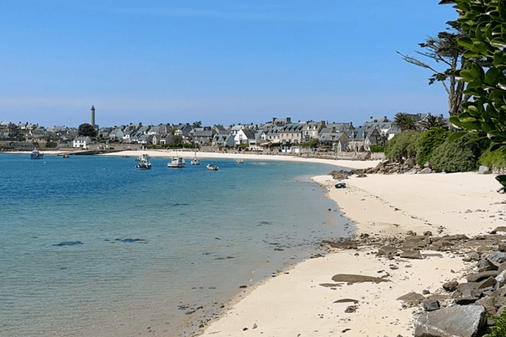 A scenic landscape view of a curved white sand beach leading toward a coastal village on Île de Batz, featuring calm turquoise water with several small moored boats, traditional stone houses along the shore, and a tall stone lighthouse visible in the distant background under a clear blue sky.