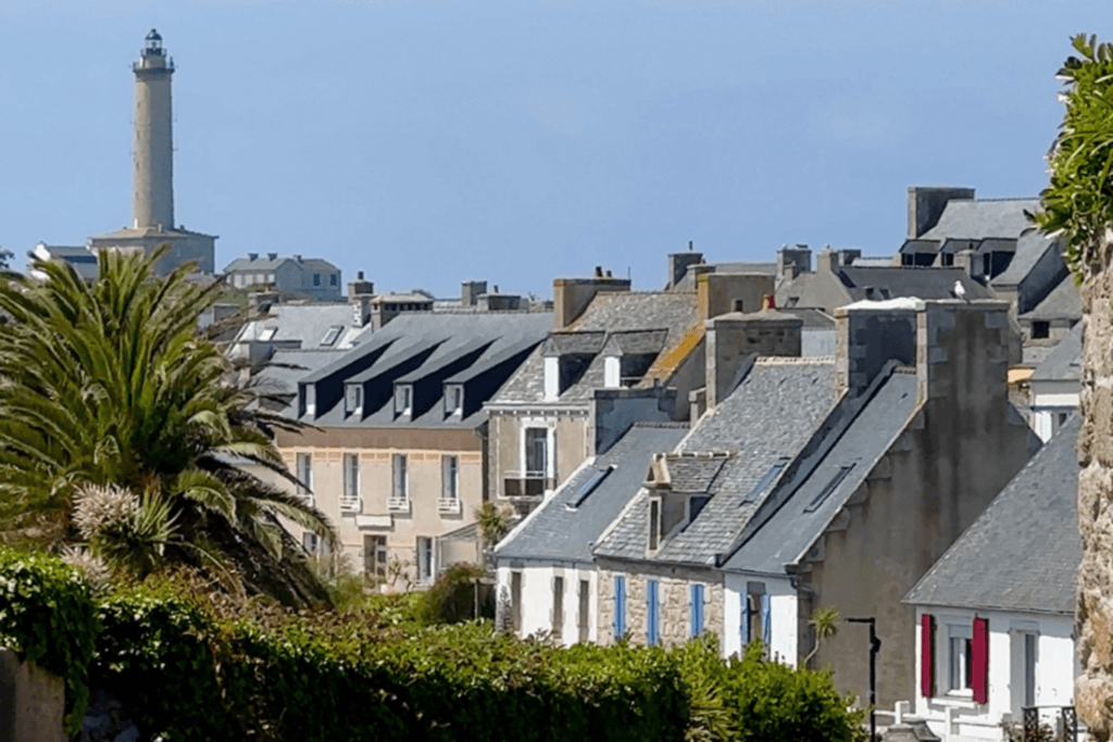 A scenic view looking across the slate-tiled rooftops and traditional stone houses of a village on Île de Batz, featuring a prominent palm tree on the left and the iconic tall stone lighthouse rising on  a hill in the background under a pale blue sky.