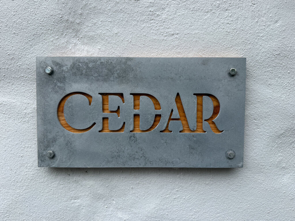 A rectangular sign mounted on a textured white wall. The name "CEDAR" is cut out of a weathered grey metal faceplate, revealing a natural wood background underneath. The plate is secured at each corner by hex bolts.