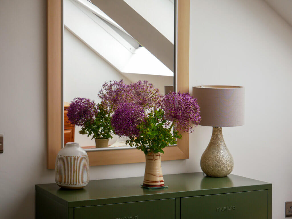 A stylized interior vignette features an olive-green metal console table situated beneath a large, wood-framed rectangular mirror. Arranged on the table's surface are three items: on the left, a textured white ceramic vase decorated with vertical brown stripes; in the centre, a slender, patterned ceramic vase holding a vibrant bouquet of large, purple allium flowers and green foliage; and on the right, a table lamp with a crackled metallic base and a cylindrical grey lampshade. The mirror behind the arrangement reflects the flowers and a bright, white-painted room with an angled ceiling and a skylight window.