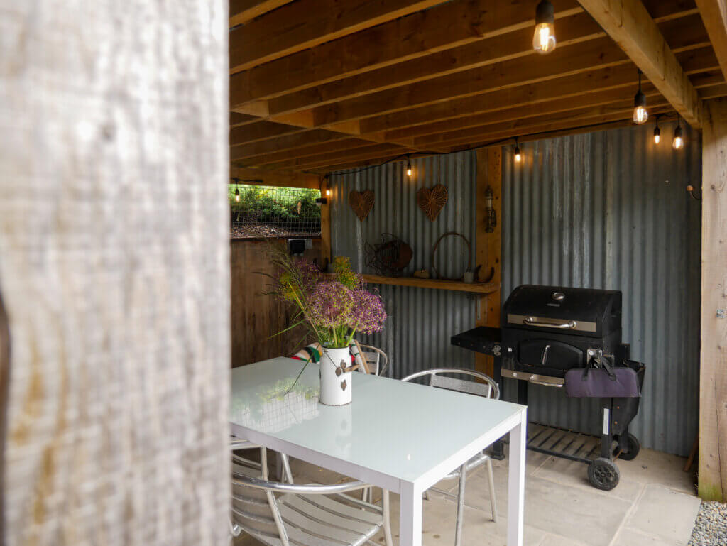 An outdoor sheltered patio area under a wooden beam roof with string lights. A white metal dining table with a glass top sits in the centre, paired with several metal chairs. In the middle of the table, a white pitcher vase holds a rustic bouquet of purple alliums and tall green grasses. The back wall is made of corrugated grey metal, decorated with woven heart shapes and a wooden shelf holding various decorative items. To the right, a black rolling barbecue grill stands against the wall.