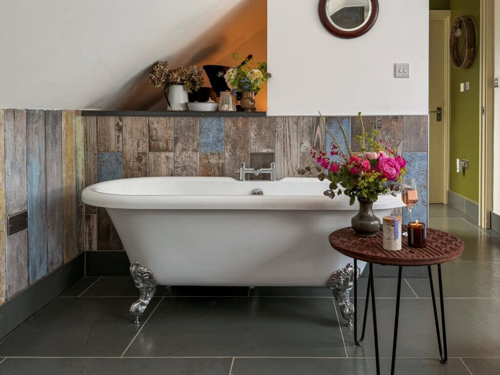 A white clawfoot bathtub with chrome ornate feet sits against a wall covered in rustic, multi-toned vertical wood planks with hints of blue, yellow, and grey. The floor is tiled with dark grey slate squares. In front of the tub, a small wooden table with black hairpin legs holds a grey vase filled with bright pink flowers, a glass jar of bath salts, a burning candle, and a glass of rose wine. The wall above the wood cladding slopes downward under an alcove where small pitchers of dried flowers are placed.