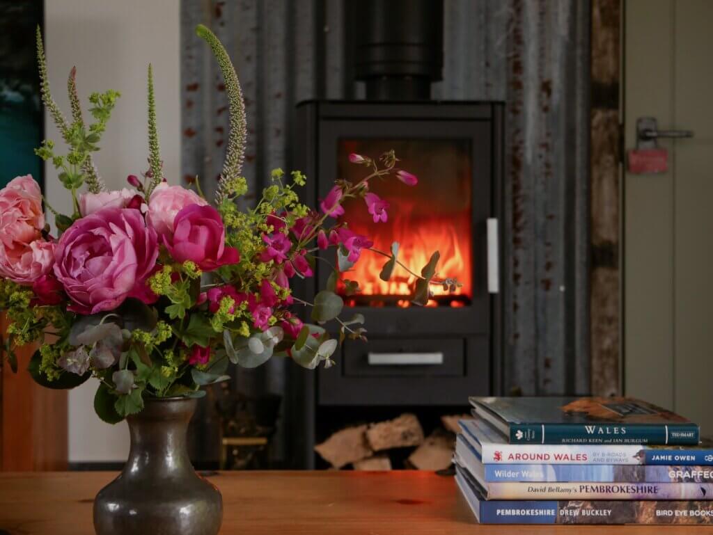 A cosy interior scene features a wooden tabletop in the foreground. On the left, a dark ceramic vase holds a lush bouquet of pink roses, magenta foxgloves, and green foliage. To the right, a stack of books includes titles like "WALES", "AROUND WALES", "Wilder Wales", "David Bellamy's PEMBROKESHIRE", and "PEMBROKESHIRE". In the soft-focused background, a black wood-burning stove glows with a bright orange fire, set against a corrugated metal wall.