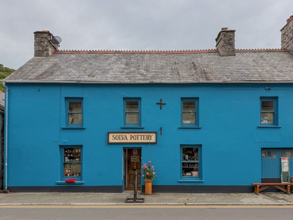 A straight-on exterior shot of "SOLVA POTTERY," a two-story building painted entirely in bright cerulean blue under a grey slate roof with two stone chimneys. A central wooden door is flanked by two large display windows filled with pottery, and a sign reading Solva Pottery hangs above the entrance. A tall terracotta pot holding pink flowers stands next to the open doorway.