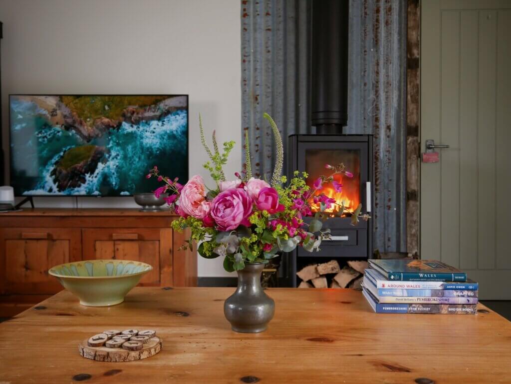 A wooden coffee table holds a vibrant bouquet of pink and purple roses in a metal vase, with a lit wood-burning stove and a TV playing a coastal scene in the background. The property is Cedar at The Longbarn a unique dog-friendly break in Wales.
