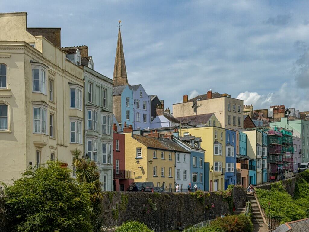 A row of colourful, multi-story seaside buildings in shades of pastel yellow, blue, pink, and cream, built along the top of a historic stone fortification wall. In the background, a tall, slender stone church spire rises against a cloudy blue sky. A stone staircase on the right leads down the ivy-covered hillside.