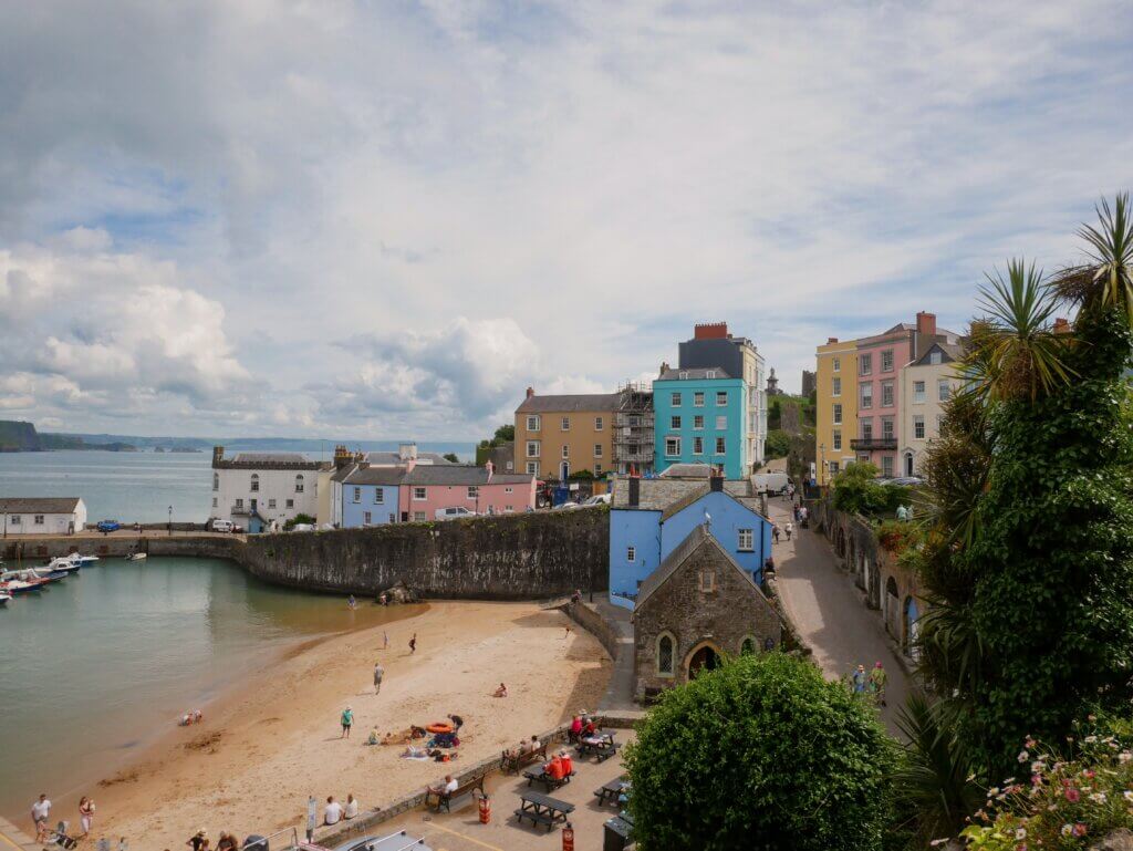 A high-angle view of a sandy beach cove nestled beside a stone harbour wall where several small boats are moored. Colourful houses stand on the cliffs overlooking the beach, and a small historic stone chapel sits next to a paved path where people are walking. On the sand and at nearby picnic tables, visitors are relaxing under a bright, partly cloudy sky.