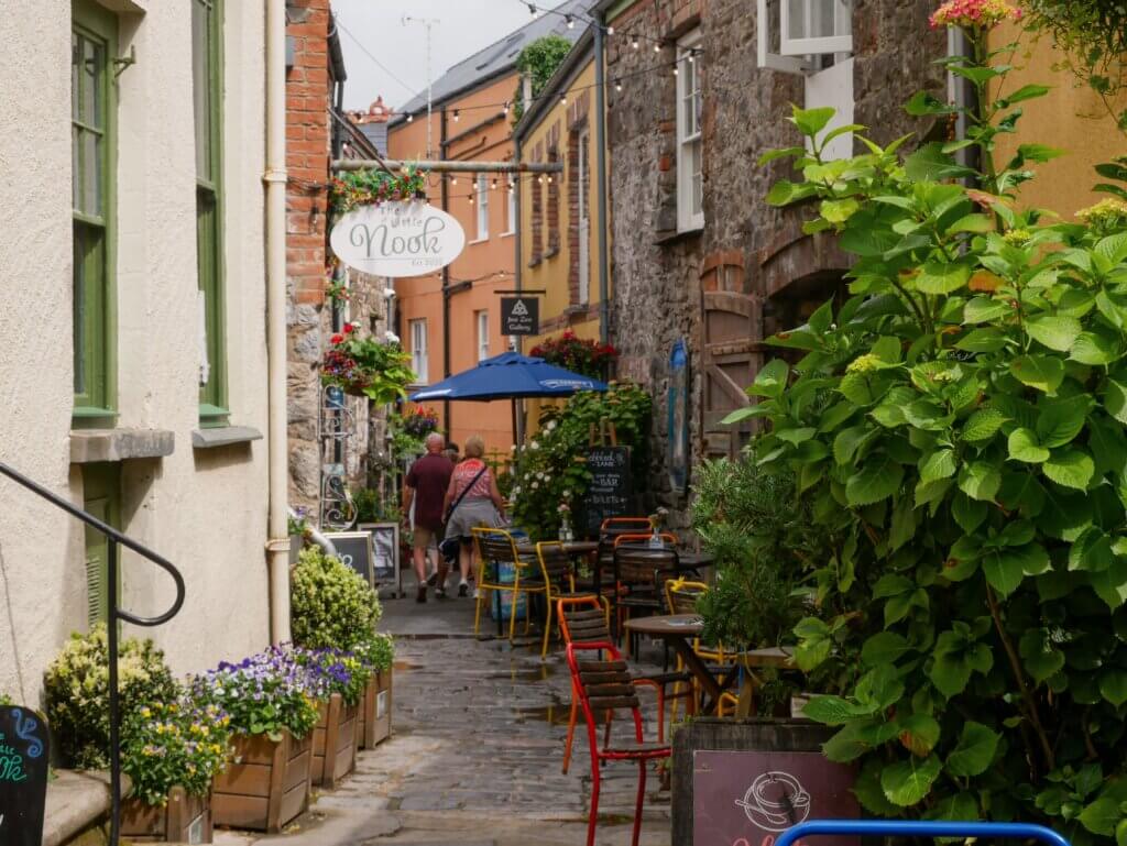 A narrow, cobblestone alleyway lined with rustic stone and brick buildings, set up with outdoor cafe seating. An oval hanging sign reads "The Little Nook Est 2025". Small wooden tables, vibrant red and yellow metal chairs, a blue patio umbrella, and abundant hanging flower baskets create an inviting atmosphere under a string of overhead lights.