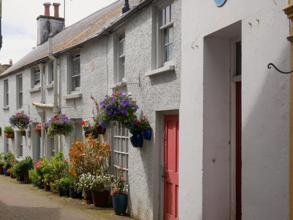 A row of traditional white-washed cottages with textured stucco walls lining a narrow paved lane. The fronts of the cottages are heavily decorated with vibrant, overflowing hanging baskets of purple and red flowers, along with numerous potted plants along the base of the walls. One cottage features a contrasting coral-pink door.