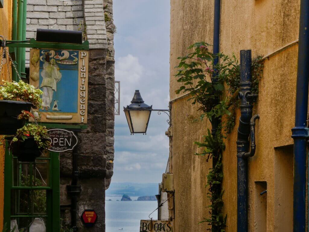 A framed view looking down a narrow space between two historic buildings toward the open sea. On the left, a green storefront features a sign reading "PLANTAGENET" and a small "OPEN" sign; on the right, an old-fashioned black street lantern hangs against a weathered yellow stucco wall with blue drainpipes and climbing green vines. In the distant blue water, small rocky islands are visible under a cloudy sky.
