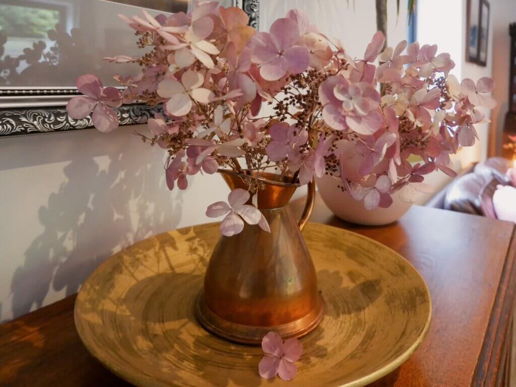 A rustic copper pitcher filled with dried pink hydrangeas sits on a matching gold-toned plate over a wooden surface.