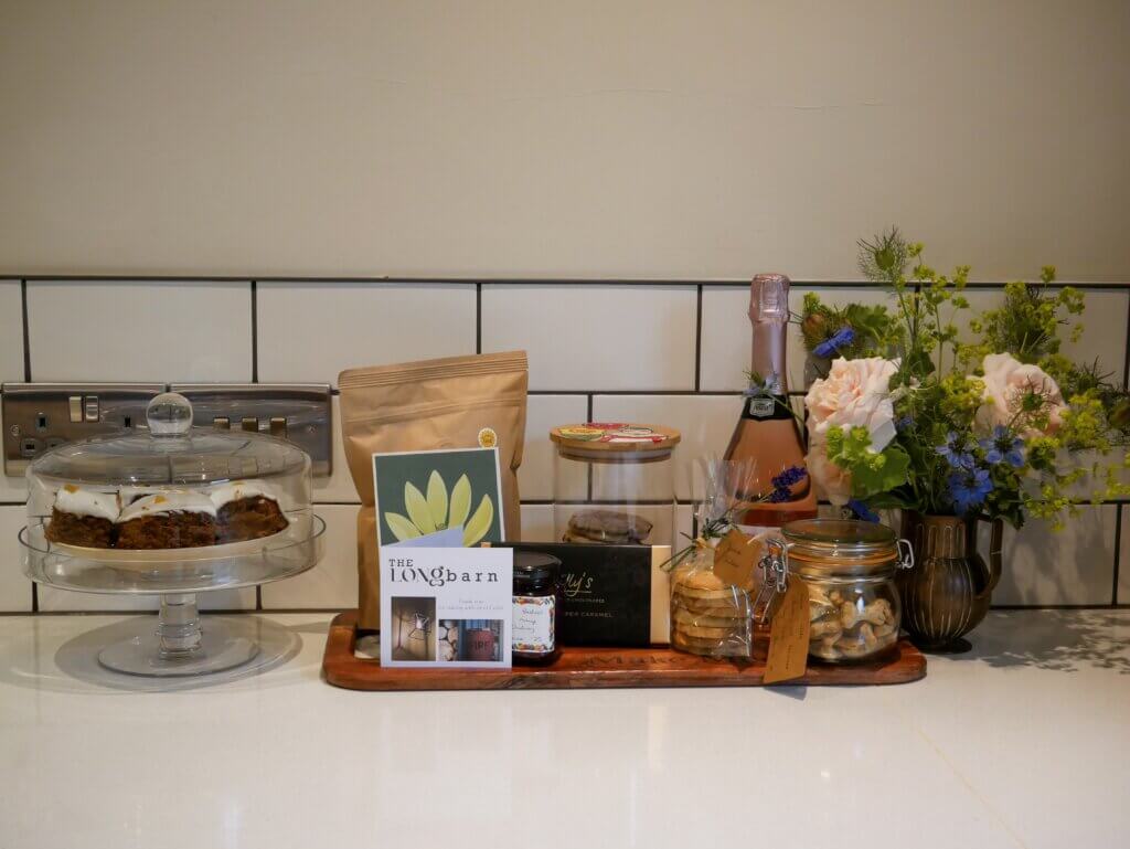 A kitchen counter features a welcome gift tray filled with wine, treats and a card from "The Longbarn", alongside a glass cake stand with frosted ginger cake slices. Cedar at The Longbarn is one of the best dog-friendly breaks in Wales.