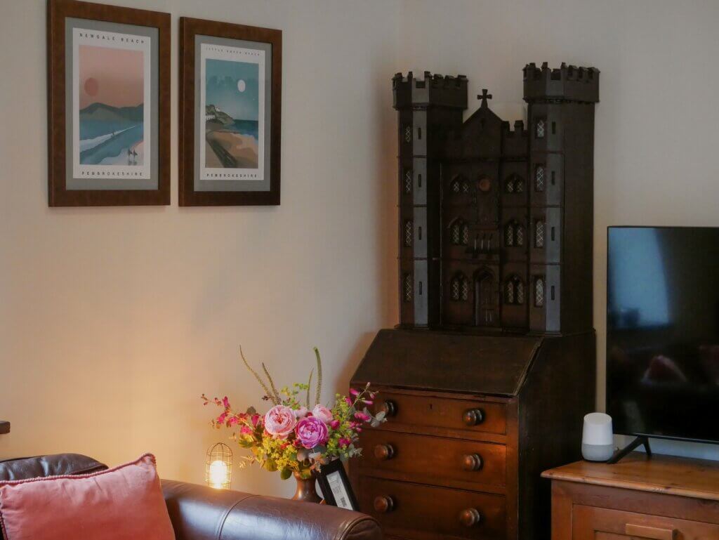 An ornate wooden bureau topped with a castle-shaped carving stands in a living room corner next to framed art and a floral arrangement.