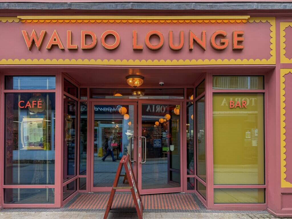 The storefront of "WALDO LOUNGE" painted in dusty rose and yellow tones with scalloped trim. The large, illuminated name sign sits above a recessed glass entryway with double doors, flanked by large windows labeled "CAFE" on the left and "BAR" on the right. A wooden A-frame chalkboard sign stands on a patterned tile floor in front of the entrance.
