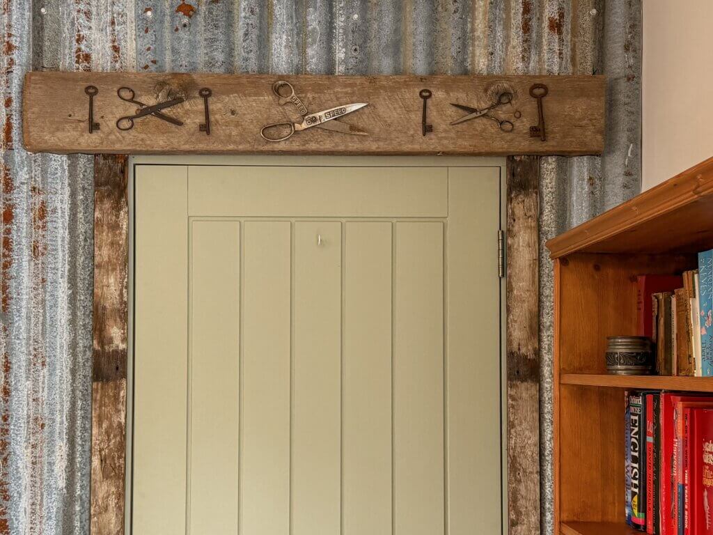 A sage green door set into a corrugated metal wall with a rustic wooden lintel above it decorated with vintage keys and scissors.