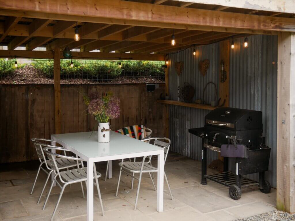 An outdoor covered patio featuring a white dining table with metal chairs, a vase of purple flowers, and a black barbecue grill against a corrugated metal wall. The space is illuminated by hanging string lights under a wooden deck ceiling.