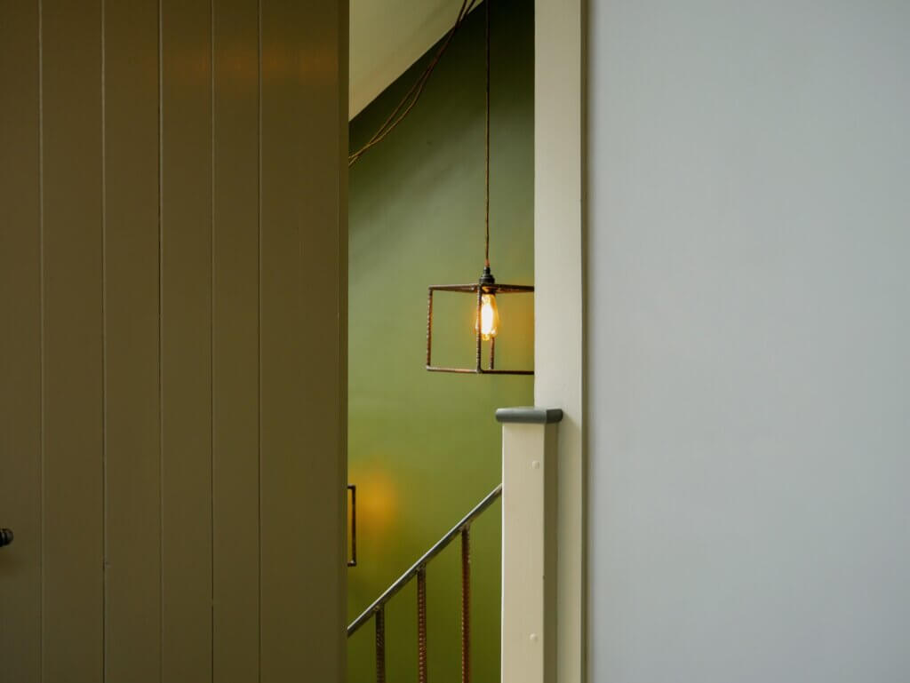 An indoor view peeking past a partially open beige paneled door into a stairwell area. A unique pendant light fixture, featuring a glowing Edison bulb enclosed in a minimalist, cubical metal rebar frame, hangs from a cord in the centre. The background reveals an olive-green painted wall and a staircase railing made of textured metal rebar. To the right of the doorway frame is a plain, light grey wall.