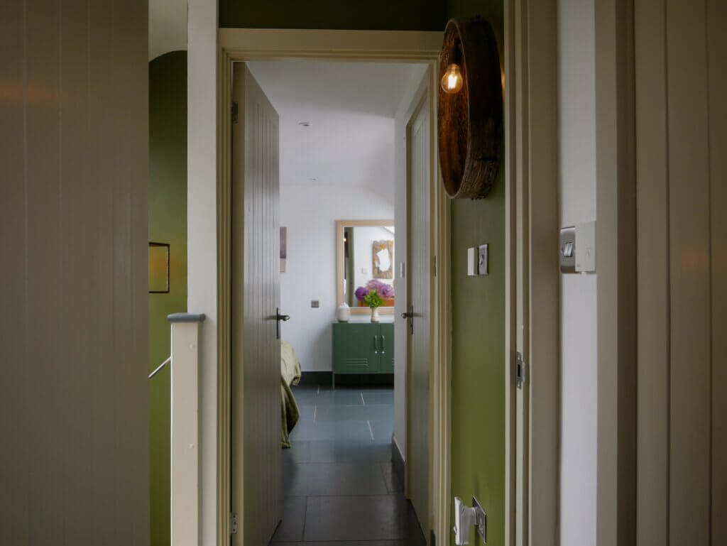 A view looking down a narrow allway with dark grey tiled flooring. The hallway walls are painted an olive green on the right, featuring a rustic wall sconce made from a circular wooden ring with an amber bulb glowing inside. An open beige paneled door in the centre leads into a brightly lit bedroom with white walls, where an olive-green metal console table, a vase of purple flowers, and a framed mirror are visible in the background. Another paneled door sits mostly closed on the right, and a glimpse of a stairwell is visible on the far left.