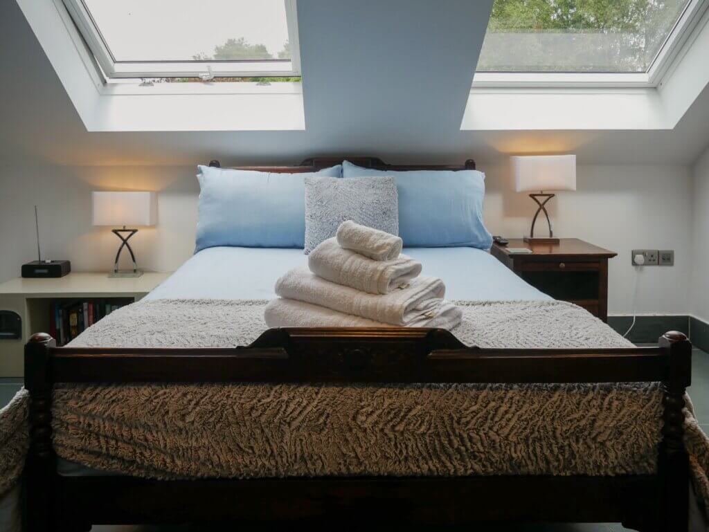 A neatly made wooden bed with light blue sheets and white towels sits directly under two large skylight windows in the attic room of Cedar at The Longbarn, the perfect dog-friendly break in Wales.
