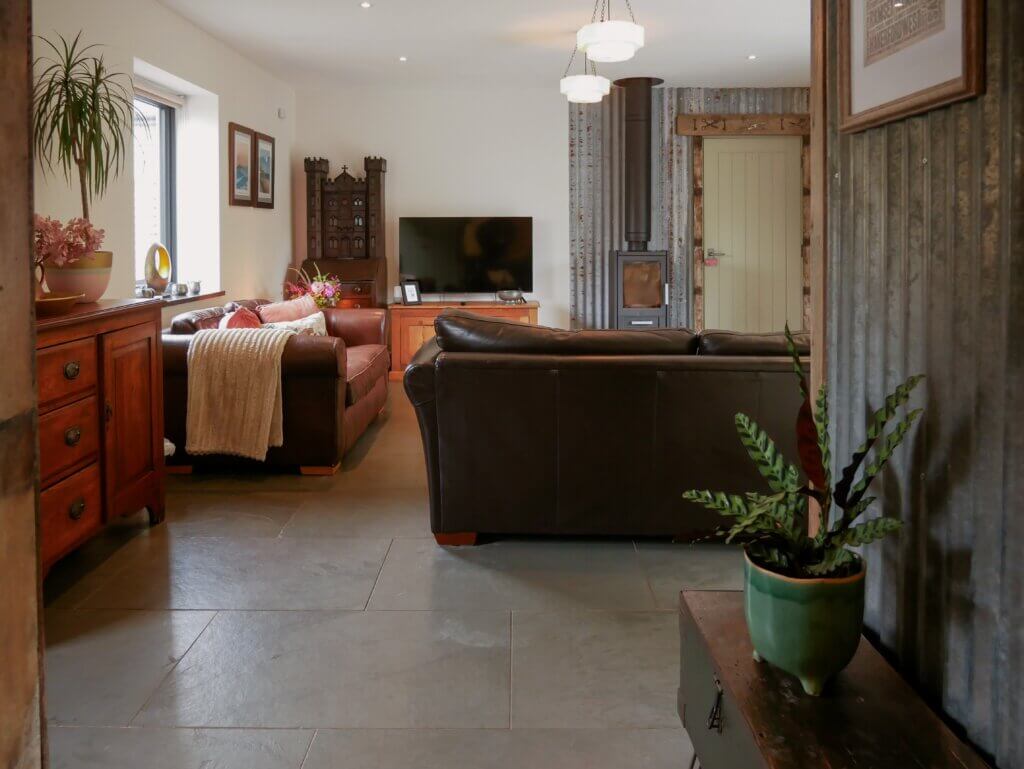A spacious, rustic-modern living room in Cedar at The Longbarn Wales, featuring brown leather sofas, slate tile flooring, a wood stove, and industrial corrugated metal accents.