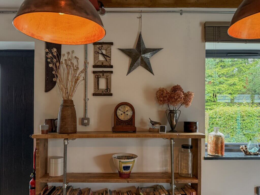 A rustic, industrial-style shelving unit made of light wood planks and silver metal pipes sits against a white wall. The top shelf displays an assortment of decor, including a tall brown textured vase with dried seed pods, an antique wooden mantle clock, a silver chalice-style vase with dried hydrangeas, and small trinkets. On the wall above the shelf hangs a large rustic metal five-point star and two small framed vintage scissor displays. Two large copper pendant lamps hang from the ceiling in the foreground, framing the scene with warm, orange light. To the right, a window looks out onto lush green trees.