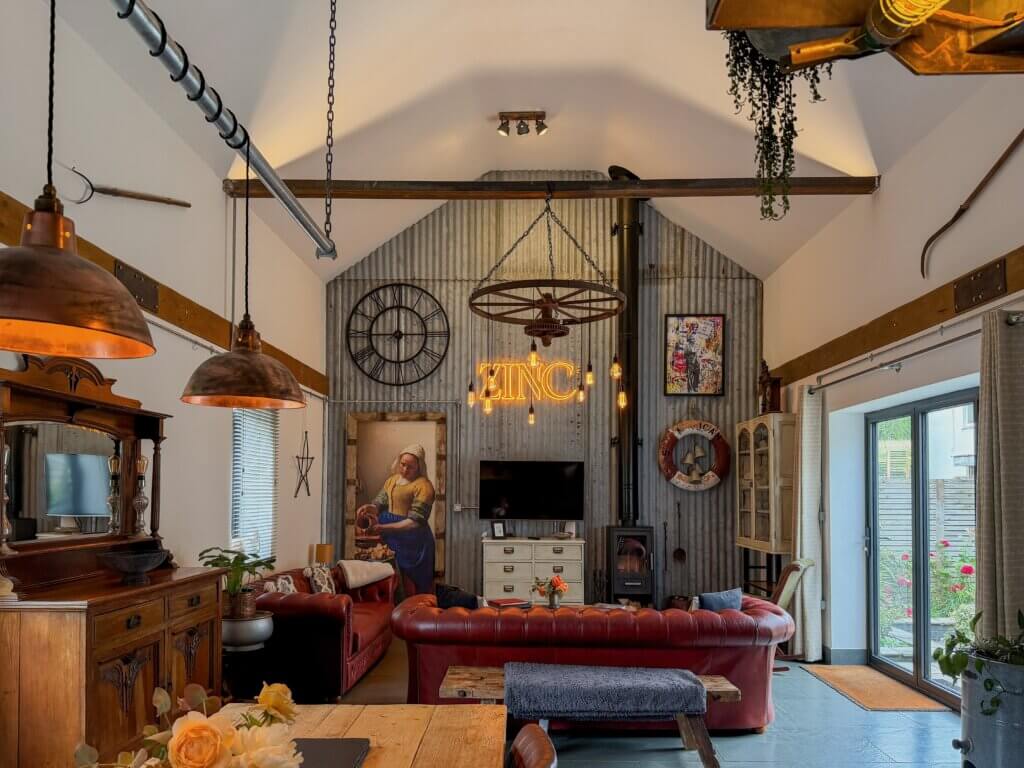 A spacious, industrial-chic living room with a vaulted white ceiling and a prominent feature wall made of vertical corrugated grey metal. In the centre of the metal wall, a glowing orange neon sign reads "ZINC" beneath a rustic wagon-wheel chandelier with hanging Edison bulbs. The room features red leather chesterfield sofas arranged around a white media console with a television. Decorative elements include a large reproduction painting of Vermeer's "The Milkmaid", a large black wall clock, a wood-burning stove, and vintage industrial lighting fixtures. Double glass doors on the right open out to a garden area.