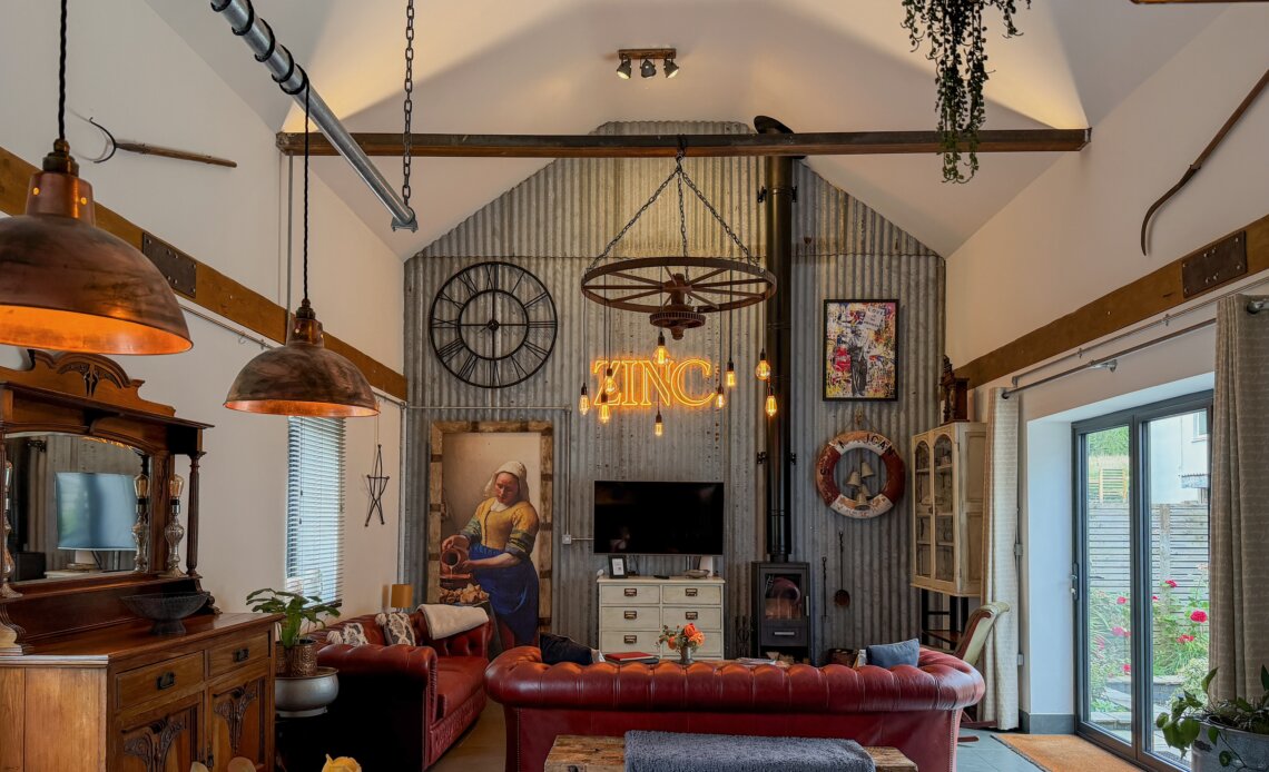 A spacious, industrial-chic living room with a vaulted white ceiling and a prominent feature wall made of vertical corrugated grey metal. In the centre of the metal wall, a glowing orange neon sign reads "ZINC" beneath a rustic wagon-wheel chandelier with hanging Edison bulbs. The room features red leather chesterfield sofas arranged around a white media console with a television. Decorative elements include a large reproduction painting of Vermeer's "The Milkmaid", a large black wall clock, a wood-burning stove, and vintage industrial lighting fixtures. Double glass doors on the right open out to a garden area.