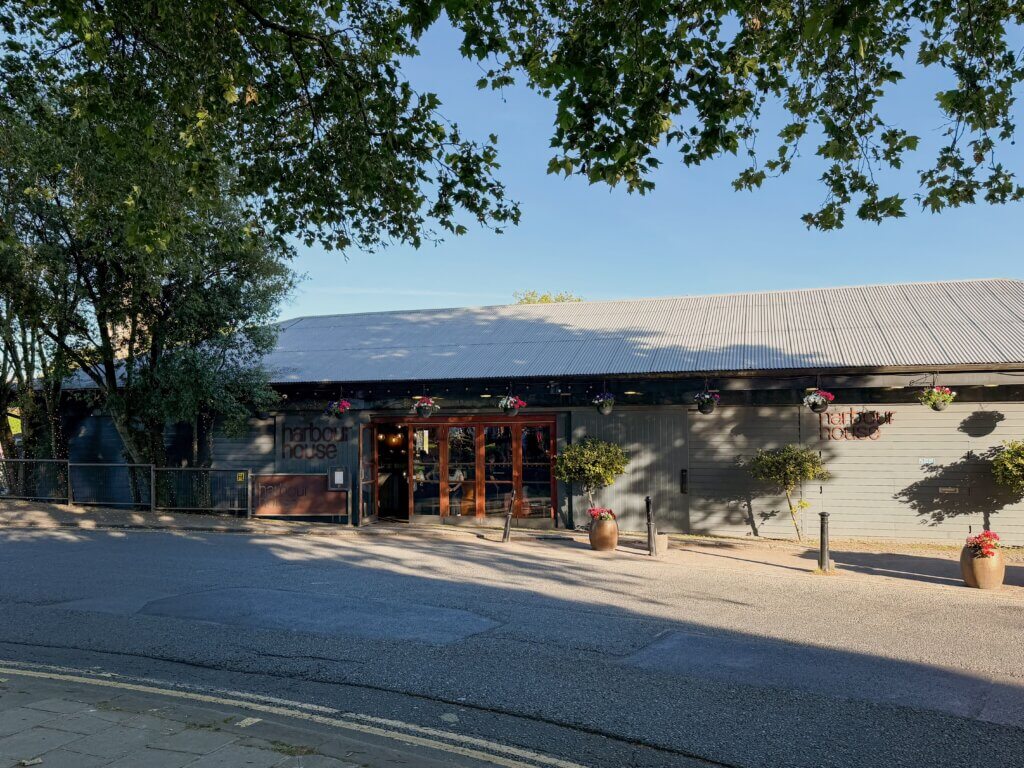 A street-level view of "Harbour House" restaurant in Bristol, a long, grey wooden building with a corrugated metal roof, large glass doors, and hanging flower baskets, shaded by overhanging tree branches.