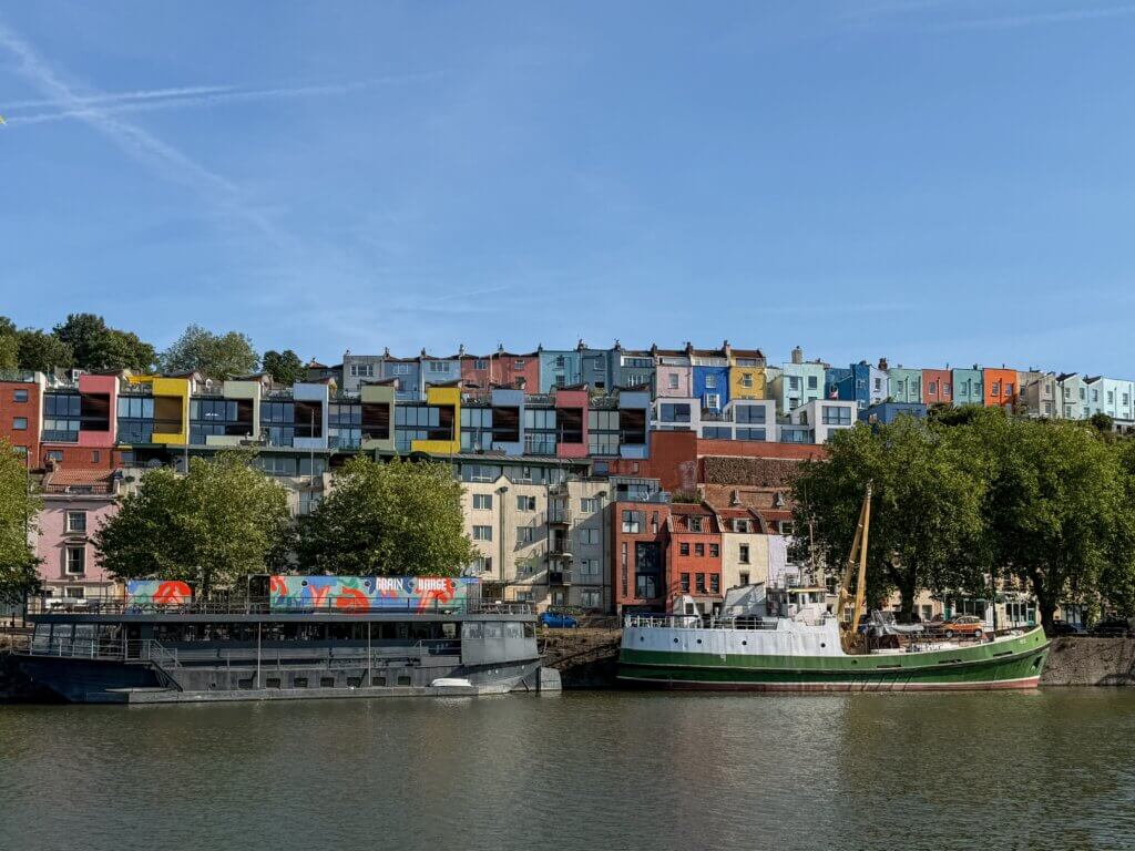 A wide shot of the river harbour in Bristol featuring a green and white ship and a dark barge labeled "Grain Barge" docked along the water, backed by rows of brightly coloured, multi-story houses rising up a hillside.