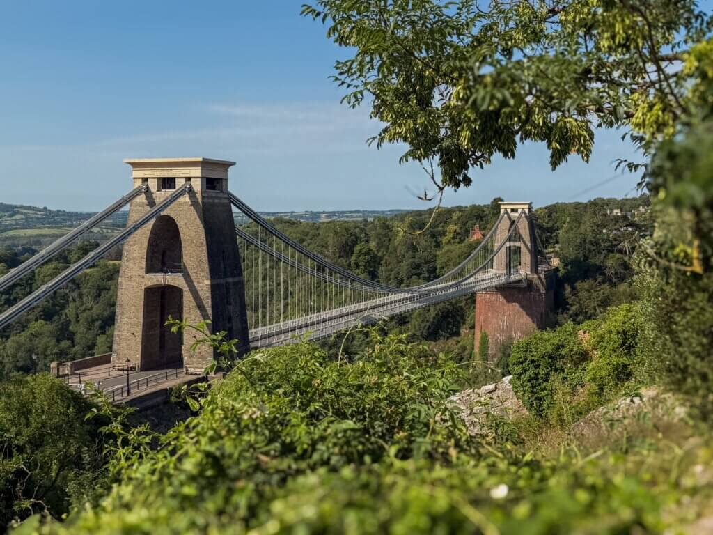 A scenic, high-angle view of the Clifton Suspension Bridge stretching across a lush green gorge under a clear blue sky, partially framed by green tree leaves in the foreground.
