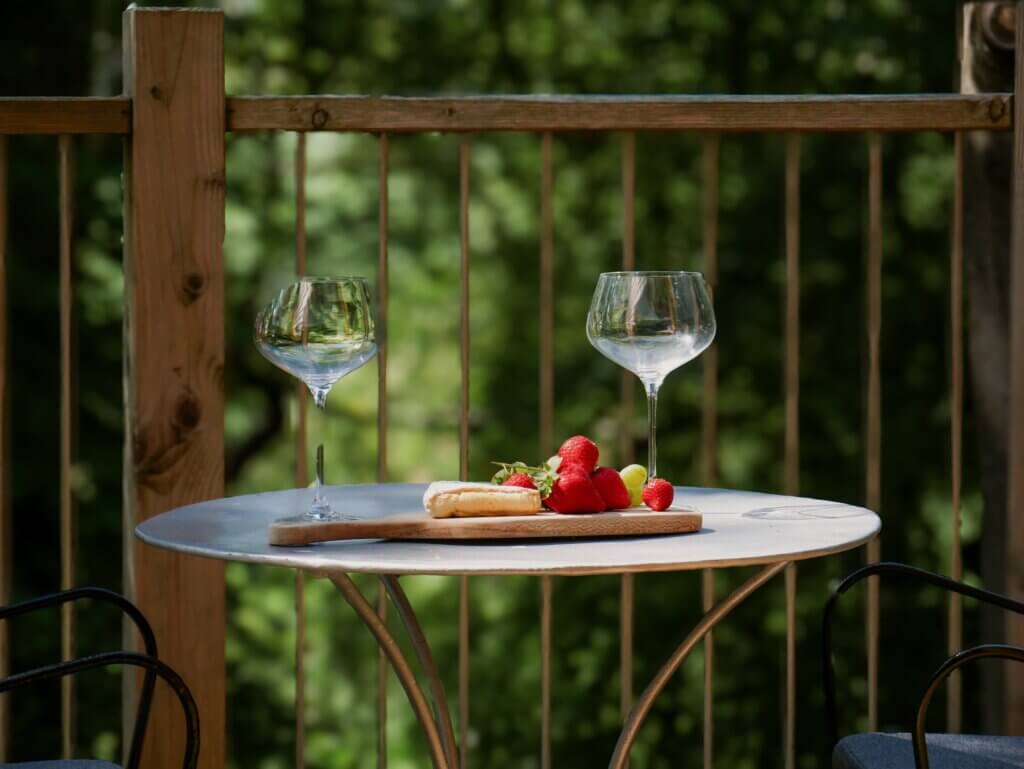 A close-up view of a round outdoor table on a deck holding two empty gin glasses and a wooden serving board topped with a wedge of cheese and fresh strawberries against a blurry green background.