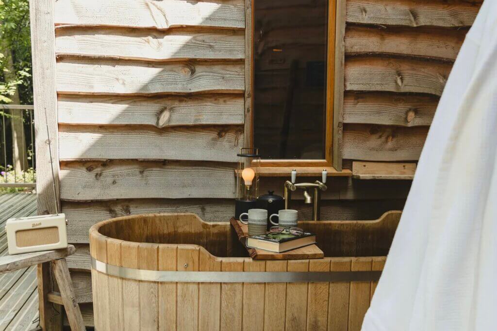 An outdoor wooden soaking tub sits on a deck next to a rustic wooden wall, with mugs, a book, and an old-fashioned radio resting nearby.