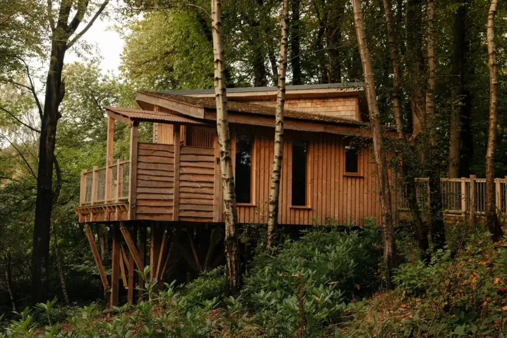 An exterior shot of a modern, rustic treehouse nestled on a sloping hillside surrounded by a dense forest.